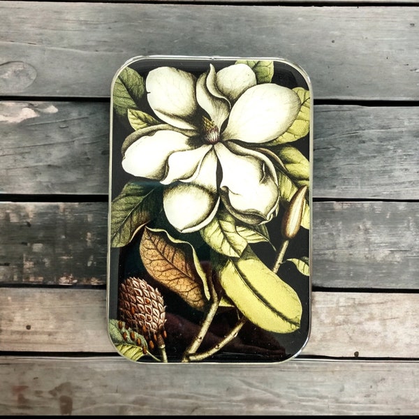 May include: Rectangular tin featuring a botanical illustration of a white magnolia flower, green leaves, and a brown seed pod on a black background. The tin has a silver border. It is set against a grey wooden surface.