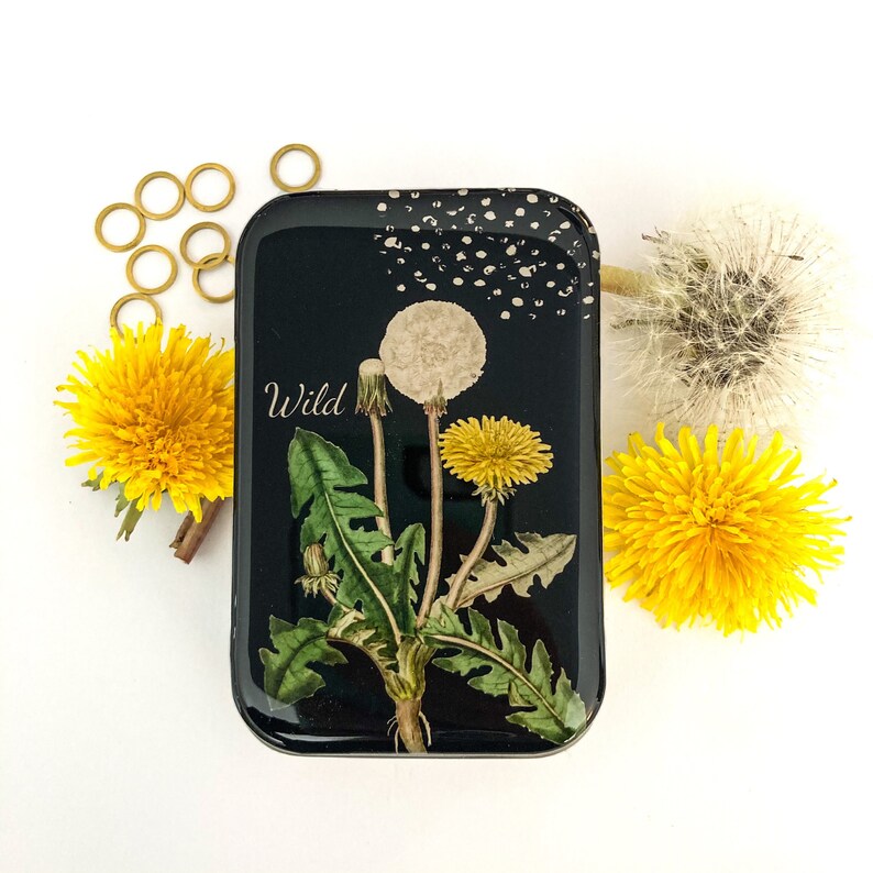 May include: A black rectangular tin with a dandelion illustration and the word "Wild" in white. The tin is surrounded by yellow dandelions and gold-colored rings on a white surface.