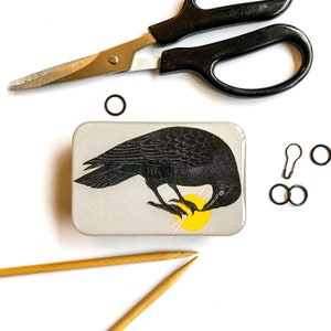 Peut inclure: Un corbeau noir avec une pelote de laine jaune dans son bec. Le corbeau est perché sur un fond gris et blanc. L'image est dans une petite boîte rectangulaire en métal.