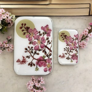 Puede incluir: Dos bandejas rectangulares con fondo blanco y bordes plateados. Las bandejas presentan un diseño floral con flores rosas y hojas verdes. Un pequeño pájaro está representado en la bandeja más grande. La bandeja más pequeña está ligeramente desplazada de la más grande.