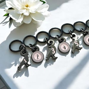 May include: A collection of black metal keychains with bird-shaped charms and numbered pink and white circular pendants. A white flower with a yellow center is in the background. The keychains are on a white surface.
