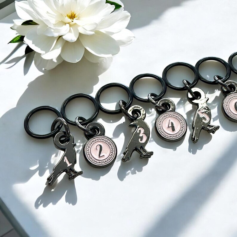 May include: A collection of black metal keychains with bird-shaped charms and numbered pink and white circular pendants. A white flower with a yellow center is in the background. The keychains are on a white surface.