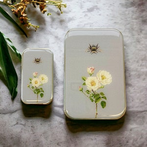 Op de afbeelding: Twee rechthoekige grijze metalen blikken met bloemen- en bijenontwerpen. De grotere blik heeft een witte roos met groene bladeren en een bij op een grijze achtergrond. De kleinere blik heeft een witte roos met groene bladeren en een bij op een grijze achtergrond.