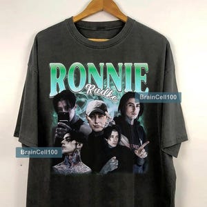 以下が含まれることがあります： 複数の人物のグラフィックと、緑と青で「Ronnie Radke」の文字がプリントされた濃いグレーのTシャツ。 ヴィンテージ感のある色あせた外観で、「BrainCell100」の文字がプリントされています。