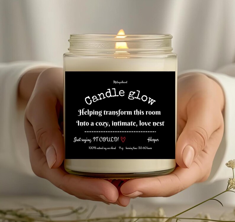 May include: A white candle with a black label that reads "Candle glow" and "Helping transform this room into a cozy, intimate, love nest." The label also includes the text "Just saying, IT COULD!" with a red heart and "Harper".