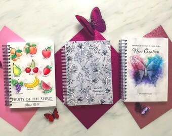 May include: Three spiral-bound notebooks with colorful covers. One features fruit illustrations and the text "Fruits of the Spirit." Another has a floral design, and the third displays a butterfly and the words "New Creation."