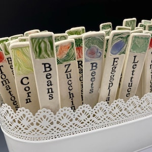 Op de afbeelding: Een verzameling keramische tuinmarkeringen met handgeschilderde illustraties en namen van verschillende groenten, waaronder tomaten, ma&iuml;s, bonen, courgette, radijs, bieten, aubergine, sla en meer.