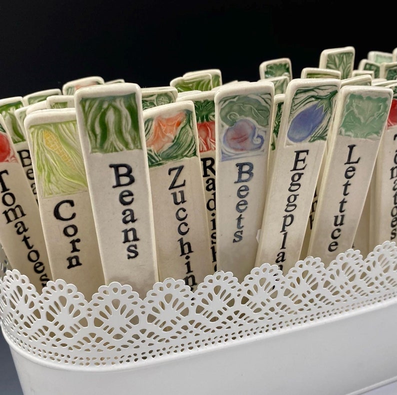 Op de afbeelding: Een verzameling keramische tuinmarkeringen met handgeschilderde illustraties en namen van verschillende groenten, waaronder tomaten, ma&iuml;s, bonen, courgette, radijs, bieten, aubergine, sla en meer.
