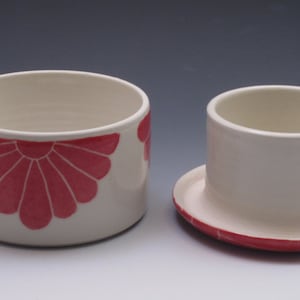 Pu&ograve; includere: Un porta burro in ceramica bianca con un motivo floreale rosa. Il porta burro ha un coperchio e una base. Il coperchio ha un piccolo pomo in cima.
