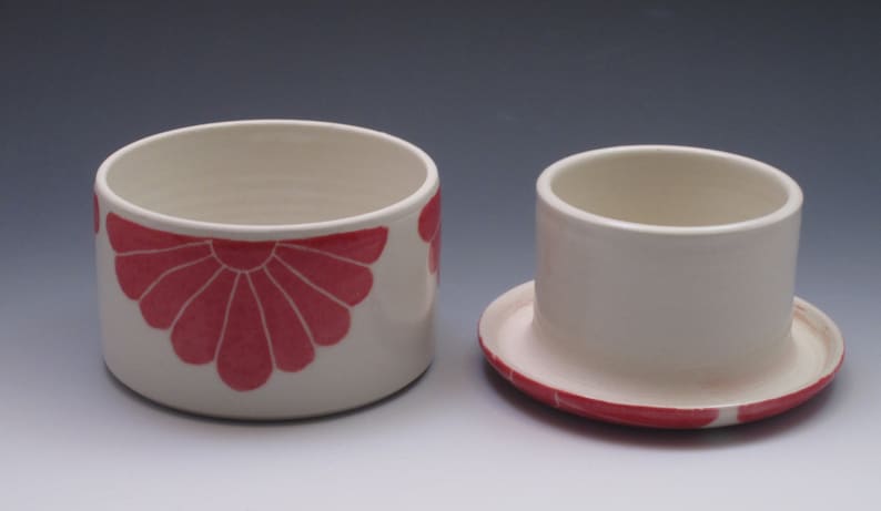 Pu&ograve; includere: Un porta burro in ceramica bianca con un motivo floreale rosa. Il porta burro ha un coperchio e una base. Il coperchio ha un piccolo pomo in cima.