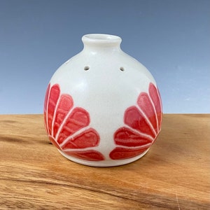 Puede incluir: Un jarrón de cerámica blanco con cuerpo redondo y una pequeña abertura en la parte superior. El jarrón está decorado con dos diseños florales rojos.