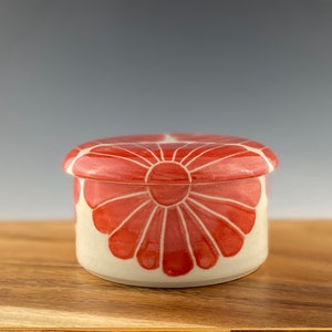 Pu&ograve; includere: Un piccolo contenitore rotondo in ceramica con un coperchio. Il contenitore &egrave; decorato con un motivo floreale rosso. Anche il coperchio &egrave; decorato con un motivo floreale rosso.