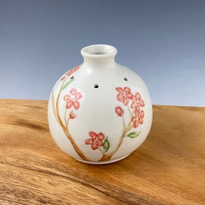 Puede incluir: Un jarrón de cerámica blanca con cuerpo redondo y una pequeña abertura en la parte superior. El jarrón está decorado con un diseño pintado a mano de flores rosas y hojas verdes.