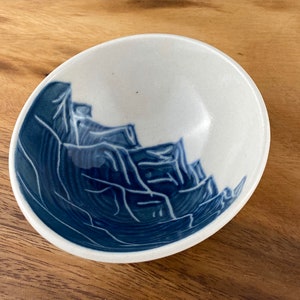 May include: A white ceramic bowl with a blue and white mountain design. The bowl is round and has a slight lip.