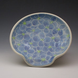 May include: A ceramic spoon rest with a light blue floral pattern. The spoon rest has a white rim and a curved indentation for holding a spoon. The flowers are light blue with green centers.