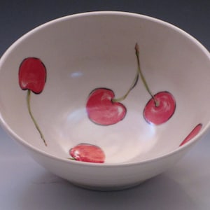 May include: White ceramic bowl with a hand-painted design of red cherries. The bowl has a rounded shape and a slightly flared rim.
