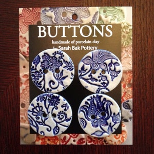 May include: Four blue and white porcelain buttons with floral designs. The buttons are handmade and say "Buttons handmade of porcelain clay by Sarah Bak Pottery".