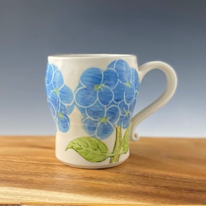 May include: White ceramic mug with a blue and green floral design. The mug has a rounded body and a handle.