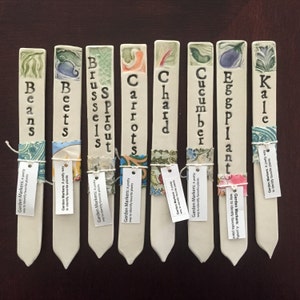 Op de afbeelding: Negen keramische tuinmarkeringen met verschillende namen en illustraties van groenten. De markeringen zijn wit met bruine letters en zijn vastgebonden met kleurrijke stoffen. De namen zijn: Bonen, Bieten, Spruitjes, Wortels, Bladspinazie, Komkommer, Aubergine, Boerenkool.