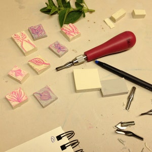 Op de afbeelding: Een close-up van een knutseltafel met verschillende gereedschappen en materialen voor het snijden en drukken. Er zijn verschillende kleine roze en witte rubberen stempels met bloemmotieven, een rood handvat snijgereedschap, een zwarte pen en een notitieboek met een spiraalbinding.
