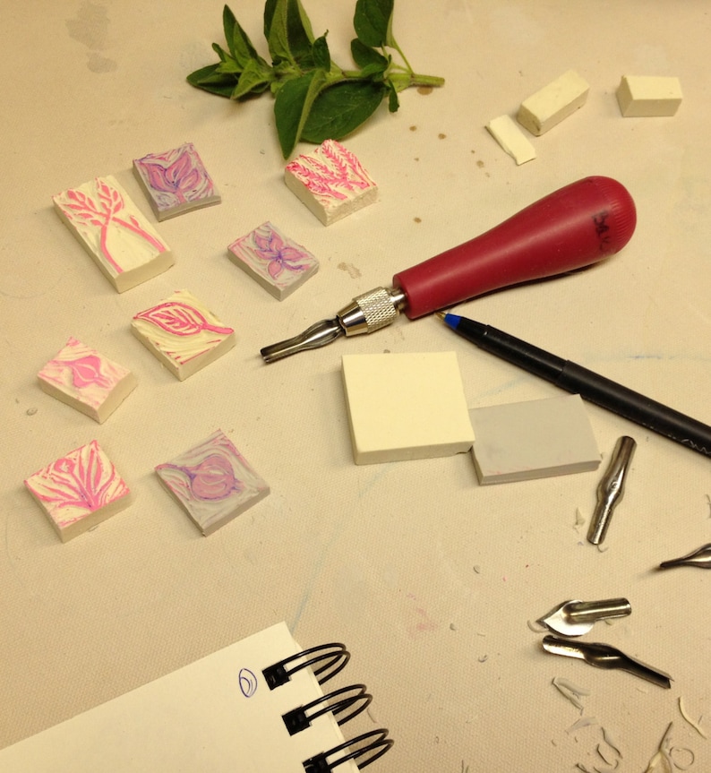 Op de afbeelding: Een close-up van een knutseltafel met verschillende gereedschappen en materialen voor het snijden en drukken. Er zijn verschillende kleine roze en witte rubberen stempels met bloemmotieven, een rood handvat snijgereedschap, een zwarte pen en een notitieboek met een spiraalbinding.