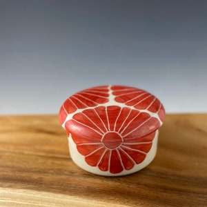 Pu&ograve; includere: Un piccolo cofanetto in ceramica bianca con un motivo floreale rosso. Il coperchio &egrave; chiuso e il cofanetto &egrave; appoggiato su una superficie di legno.