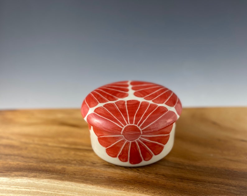 Pu&ograve; includere: Un piccolo cofanetto in ceramica bianca con un motivo floreale rosso. Il coperchio &egrave; chiuso e il cofanetto &egrave; appoggiato su una superficie di legno.
