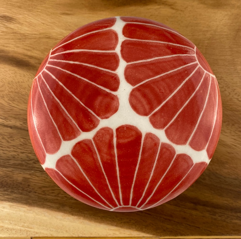 Pu&ograve; includere: Una scatola rotonda in ceramica rossa con un design geometrico bianco. Il design presenta una serie di forme a ventaglio sovrapposte.
