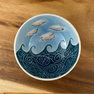 May include: A white ceramic bowl with a blue and white design of waves and clouds. The waves are outlined in black and have a swirling pattern. The clouds are pink and white.