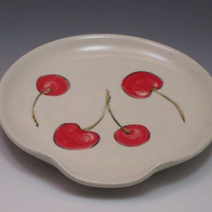Puede incluir: Apoyacucharas de cerámica blanquecina con un diseño pintado a mano de cuatro cerezas rojas. Las cerezas tienen tallos verdes y están delineadas en negro. El apoyacucharas tiene una forma curva con una pequeña hendidura.