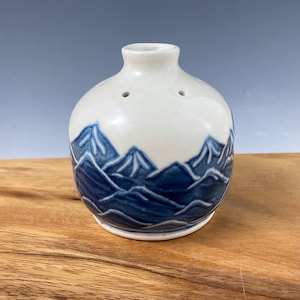 Puede incluir: Un jarrón de cerámica blanco con un diseño de montaña azul. El jarrón tiene un cuerpo redondo y un cuello estrecho. Hay pequeños agujeros en el cuello del jarrón.