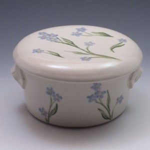 Puede incluir: Un recipiente de cerámica blanca con tapa, que presenta un delicado diseño floral de flores azules de nomeolvides y tallos verdes. El recipiente tiene dos pequeñas asas a los lados.