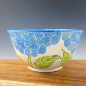 May include: A white ceramic bowl with a blue and green floral design. The bowl is decorated with blue hydrangeas and green leaves.