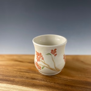 May include: A white ceramic cup with a floral design. The cup has a rounded bottom and a flared rim. The floral design features a branch with orange flowers.