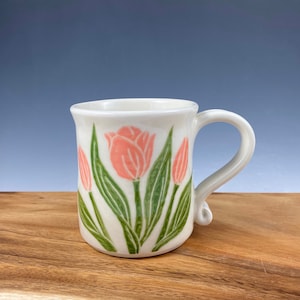 Puede incluir: Taza de cerámica blanca con asa, decorada con un diseño de tulipanes rosas y hojas verdes. La taza tiene un borde ligeramente acampanado y un asa curvada. El fondo es un degradado de azul y la taza está sobre una superficie de madera.