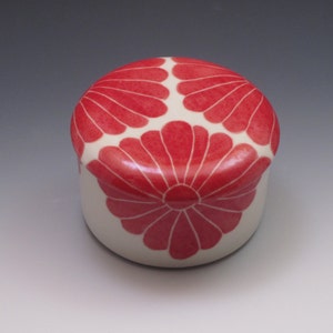 Pu&ograve; includere: Una piccola scatola rotonda in ceramica con una base bianca e un motivo floreale rosso sul coperchio. Il coperchio presenta tre fiori stilizzati con un cerchio centrale.