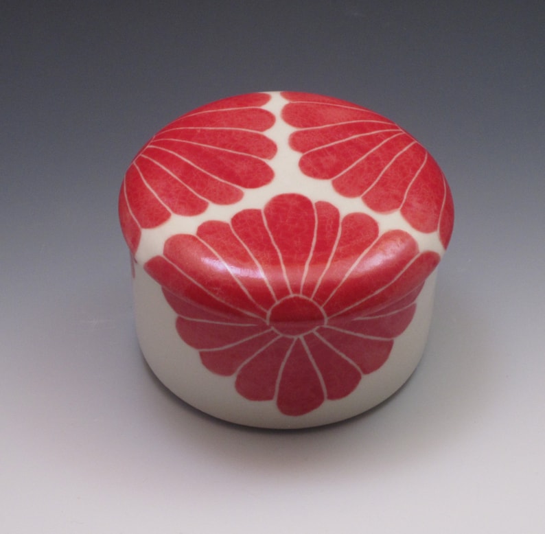 Pu&ograve; includere: Una piccola scatola rotonda in ceramica con una base bianca e un motivo floreale rosso sul coperchio. Il coperchio presenta tre fiori stilizzati con un cerchio centrale.