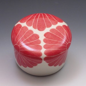 Pu&ograve; includere: Una scatola portaoggetti in ceramica bianca con un coperchio. Il coperchio &egrave; decorato con tre motivi floreali stilizzati rossi.