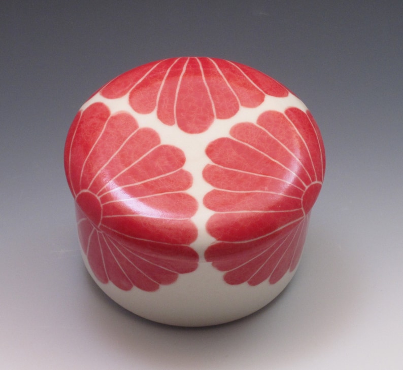 Pu&ograve; includere: Una scatola portaoggetti in ceramica bianca con un coperchio. Il coperchio &egrave; decorato con tre motivi floreali stilizzati rossi.