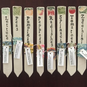 Op de afbeelding: Set van tien keramische tuinmarkeringen met kleurrijke bloemenmotieven en handgeschreven namen van groenten: Sla, Erwten, Pompoen, Paprika, Radijs, Spinazie, Tomaten, Courgette. Elke marker heeft een klein label met de tekst "Garden Markers: A pretty way to identify favorite plants".