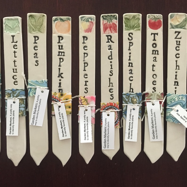 Op de afbeelding: Set van tien keramische tuinmarkeringen met kleurrijke bloemenmotieven en handgeschreven namen van groenten: Sla, Erwten, Pompoen, Paprika, Radijs, Spinazie, Tomaten, Courgette. Elke marker heeft een klein label met de tekst "Garden Markers: A pretty way to identify favorite plants".