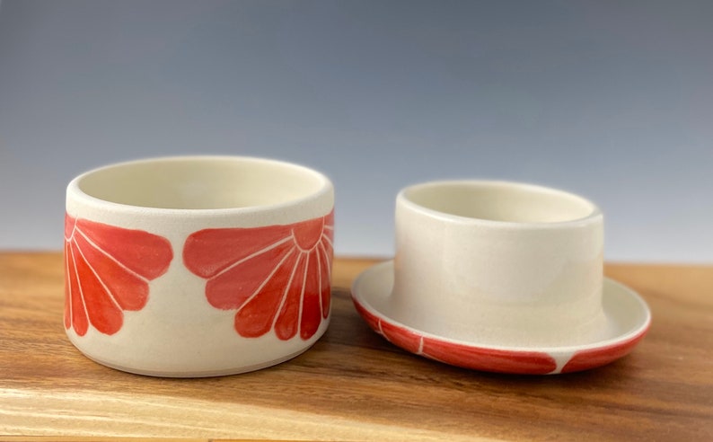 Pu&ograve; includere: Un porta burro in ceramica bianca con un motivo floreale rosso. Il porta burro ha un coperchio e una base. La base ha un motivo floreale rosso attorno al bordo.