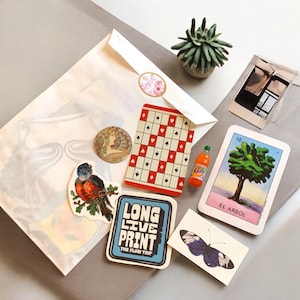 May include: A collection of stickers, cards, and a small Fanta bottle arranged on a gray surface. Items include a playing card with red and black patterns, a sticker with a bird on a branch, and a sticker with the text "LONG LIVE PRINT THE FLOW TRIP".