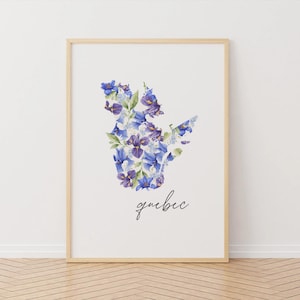 Peut inclure: Une affiche encadrée représentant la forme du Québec, remplie de fleurs à l'aquarelle dans des tons de bleu et de violet. Le mot "quebec" est écrit en cursive sous le motif floral. L'œuvre est présentée dans un cadre en bois clair.