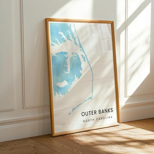 Puede incluir: Una impresión artística enmarcada que presenta un mapa de Outer Banks, Carolina del Norte. El mapa es en tonos azules y blancos, sobre un fondo blanco. La impresión está en un marco de madera y se apoya contra una pared blanca y un suelo de madera.
