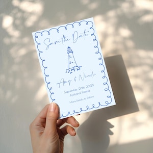 Peut inclure: Carte Save the Date bleu clair avec une illustration de phare bleu marine et du texte. La carte comprend les noms "Amy & Nicole", la date "26 septembre 2026" et le lieu "Portland, Maine". La carte a une bordure décorative.