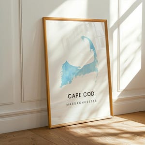 Op de afbeelding: Een ingelijste print van Cape Cod, Massachusetts, in een lichtblauwe aquarel stijl. De print zit in een natuurlijke houten lijst en leunt tegen een witte muur. De tekst "CAPE COD MASSACHUSETTS" is in het zwart gedrukt.