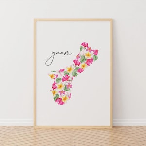 May include: A framed print featuring the outline of Guam filled with watercolor flowers in shades of pink, yellow, and green. The word "guam" is written in cursive above the floral design. The frame is light brown.