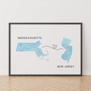 Puede incluir: Lámina enmarcada con mapas en acuarela de Massachusetts y Nueva Jersey, conectados por una línea en forma de corazón. Las palabras "Massachusetts" y "New Jersey" están impresas encima y debajo de los respectivos estados. La obra está en un marco negro.