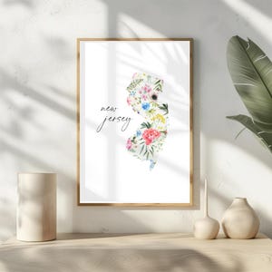 May include: A white framed print with a floral design of the state of New Jersey. The text "new jersey" is written in black cursive script.